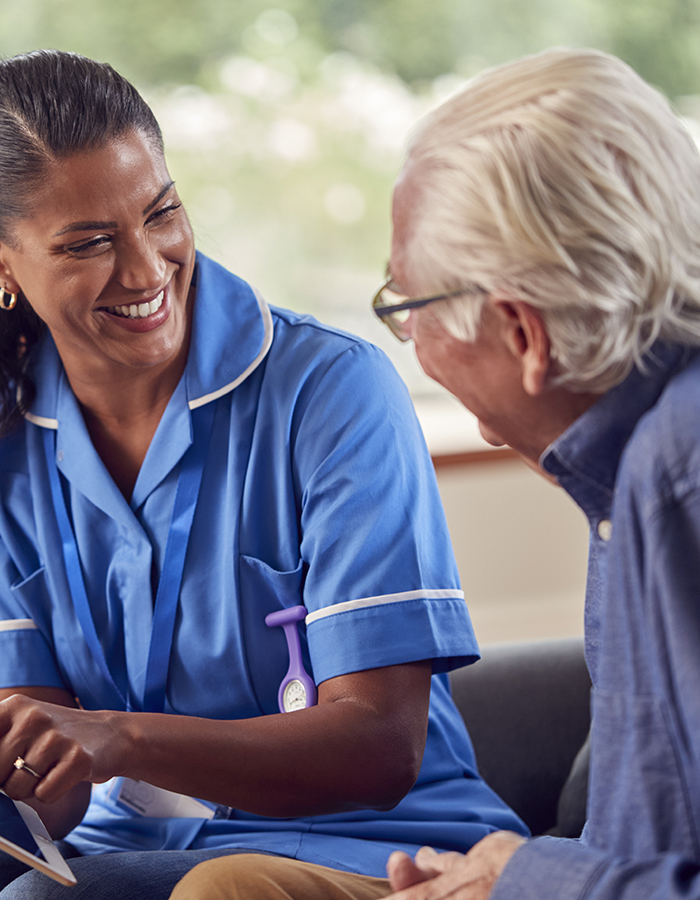 Senior Couple At Home With Man Talking To Female Nurse Or Care Worker Using Digital Tablet