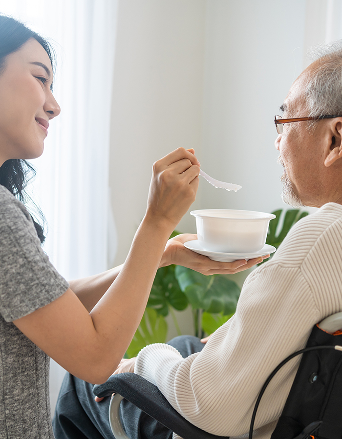 Asian beautiful young daughter support to elderly father on wheelchair. Attractive woman serving and feeding food to older patient grandfather in living room at home. Family Healthcare medical concept